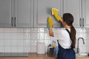 A woman wearing yellow gloves and an apron cleans a gray kitchen cabinet with a spray bottle and cloth, showcasing professional airbnb cleaning skills.