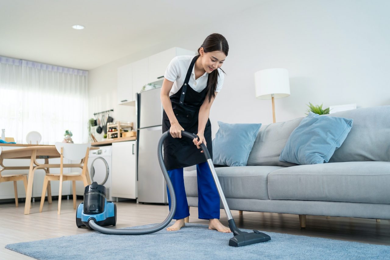 A woman wearing an apron vacuums the floor in a modern living room with a gray sofa and blue cushions, showcasing professional house cleaning.