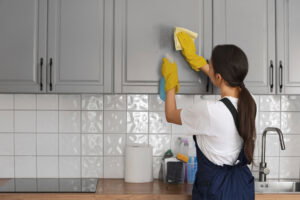 A person wearing yellow gloves cleans a gray kitchen cabinet with a cloth and spray bottle, providing deep cleaning worthy of a professional cleaning company.