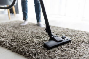 A cleaning lady is vacuuming a thick, light brown carpet with a black vacuum cleaner, her legs and white socks visible in the background—perfect for thorough move out cleaning. Carpet cleaning