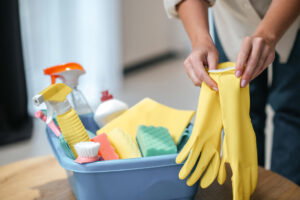 Preparing for weekly cleaning, a person puts on yellow gloves beside a bin filled with cleaning supplies and sponges.